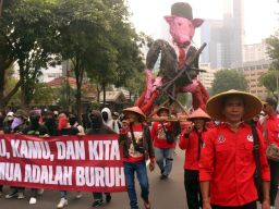 Massa aksi membawa replika patung babi berdasi lengkap dengan senapan, serta membentangkan spanduk berisi tuntutan para buruh.